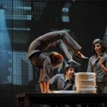 Szene aus der Show Cirkopolis: Frau macht Handstand auf einem Tisch mit einem Stapel Unterlagen. Im Hintergrund zwei weitere Artisten und eine Industriehalle als Kulisse