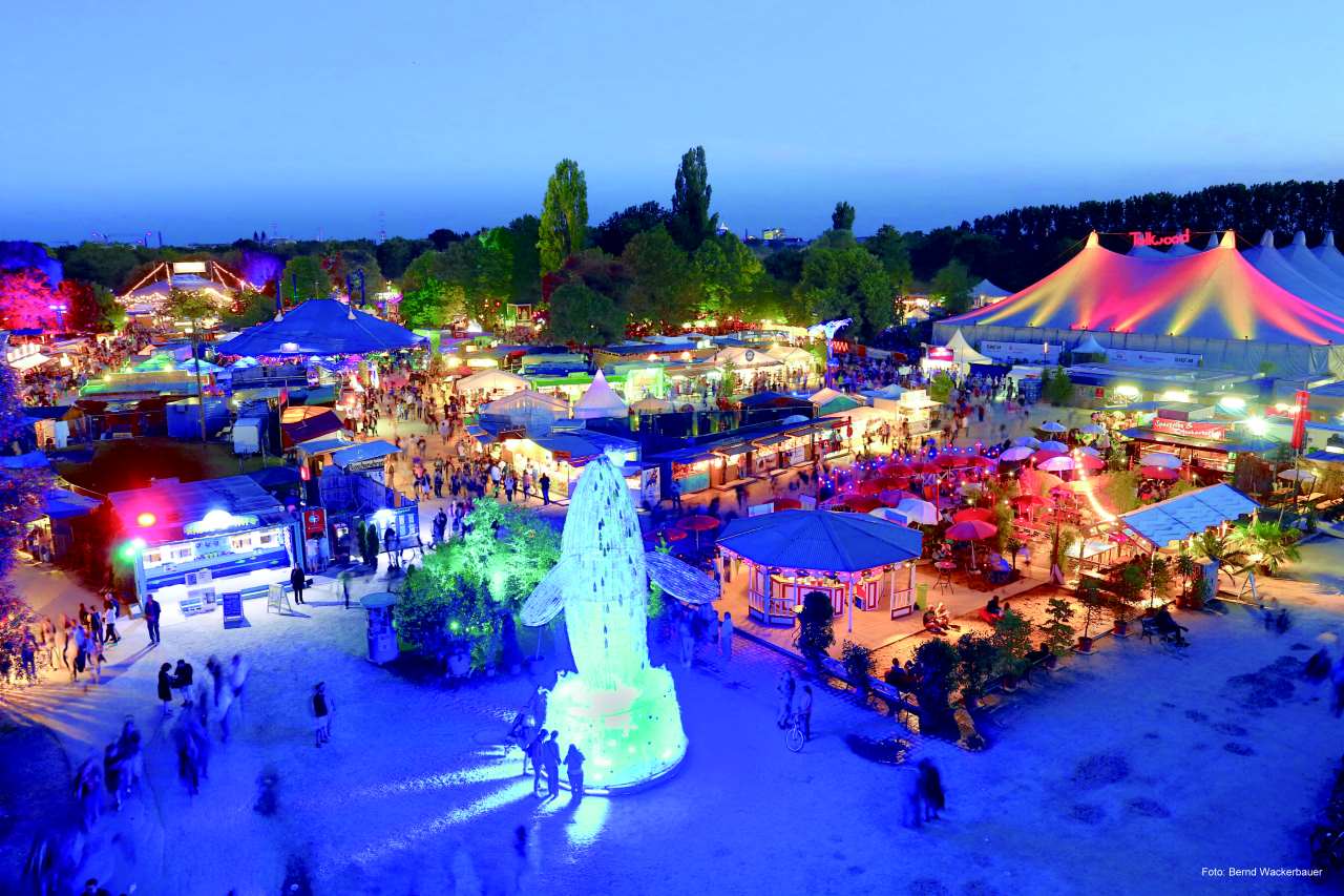 Blick über das beleuchtete Tollwood Sommerfestival. Im Vordergrund ein blaubeleuchteter Waal, der scheinbar aus dem Boden auftaucht.