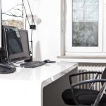 Weißer Schreibtisch mit Monitor und Telefonen sowie einem Drehstuhl. Im Hintergrund ein Fenster