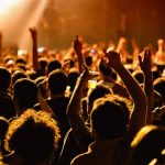 Konzertpublikum vor eine Bühne. Die Besucher trinken Bier und halten die Arme hoch.