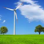 Windrad auf einer grünen Wiese mit zwei Bäumen. Der Himmel ist blau mit weißen Wolken.