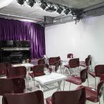 Kleinkunstbühne mit Klavier und einem purpurfarbenen Vorhang. Davorstehen einzelne moderne Stühle mit kleinen Beistelltischen