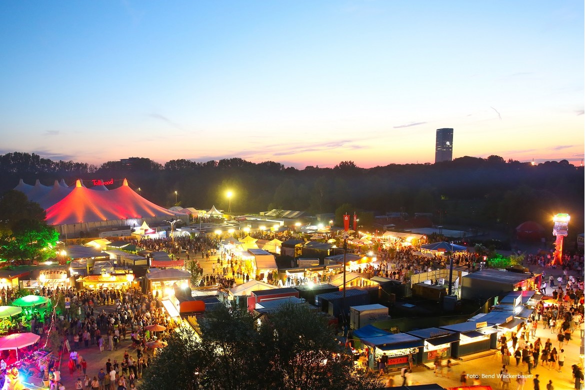 Ein Blick vom Olympiaberg über das festlich beleuchtete Tollwood Sommerfestival hinüber zum o2 Hochhaus