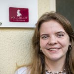 zu sehen ist Tabea Kraus im Vordergrund von dem Logo vollblut LiveMarketing. Sie hat ein breites Lächeln aufgesetzt, trägt Schmuck und ein weißes Oberteil, dass man nur zur Hälfte sieht den nur ihr Hals und Kopf ist zu sehen