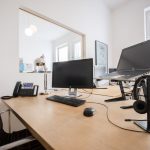 zu sehen ist ein Arbeitsplatz der modern Ausgestattet ist mit einem Laptop, Computer, Telefon, Headset, Tastatur, Maus und einer Lampe. Im Hintergrund sind Fenster zu sehen