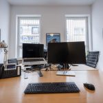 zu sehen ist ein Arbeitsplatz der modern Ausgestattet ist mit einem Laptop, Computer, Telefon, Tastatur, Maus und einer Lampe. Im Hintergrund sieht man Bilder an der Wand hängen und es ist schön hell im Raum, man sieht zwei Fenster