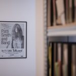 zu sehen ist ein eingerahmtes Plakat von Patti Smith and Band mit originaler Unterschrift, das Bild ist eingerahmt und hängt an einer weißen Wand, auf dem Foto ist links verschwommen ein Regal mit Ordnern zu sehen