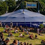 Servicekräfte für Seniorennachmittag - Das Bild zeigt das Anechser Zelt auf dem Tollwood Sommerfestival. Auf dem grünem Hügel vor dem Zelt genießen zahlreiche Besucher*innen die Sonne.