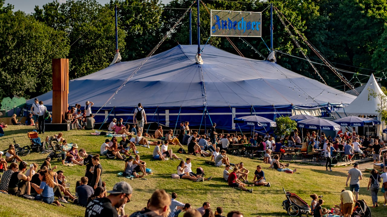 Servicekräfte für Seniorennachmittag - Das Bild zeigt das Anechser Zelt auf dem Tollwood Sommerfestival. Auf dem grünem Hügel vor dem Zelt genießen zahlreiche Besucher*innen die Sonne.