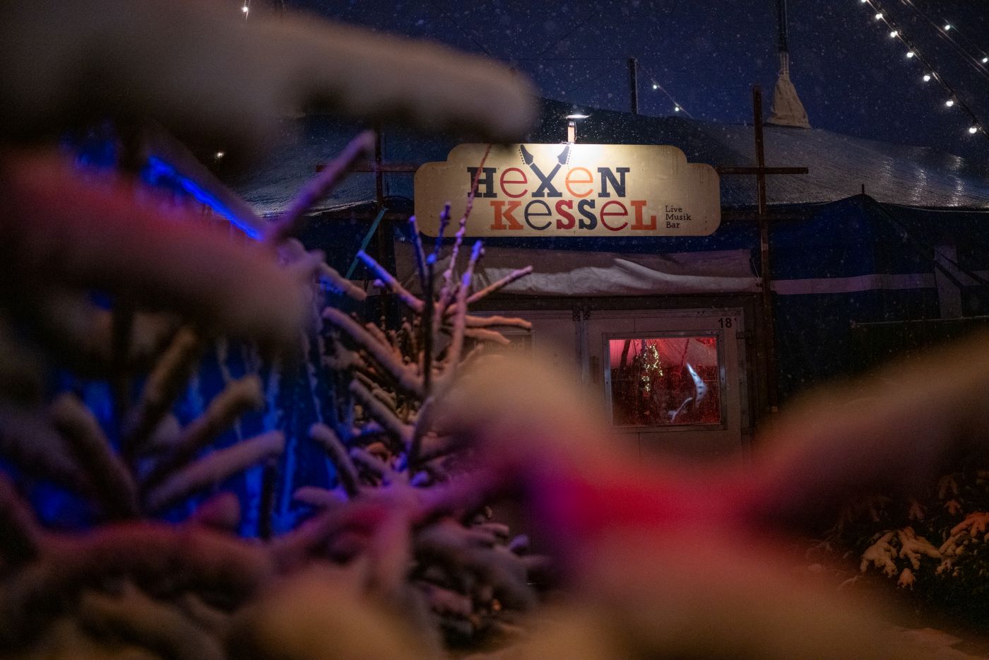 Weihnachtsfeiern auf dem Tollwood Winterfestival: Das Bild zeigt den Hexenkessel durch verschneite Tannenbäume.